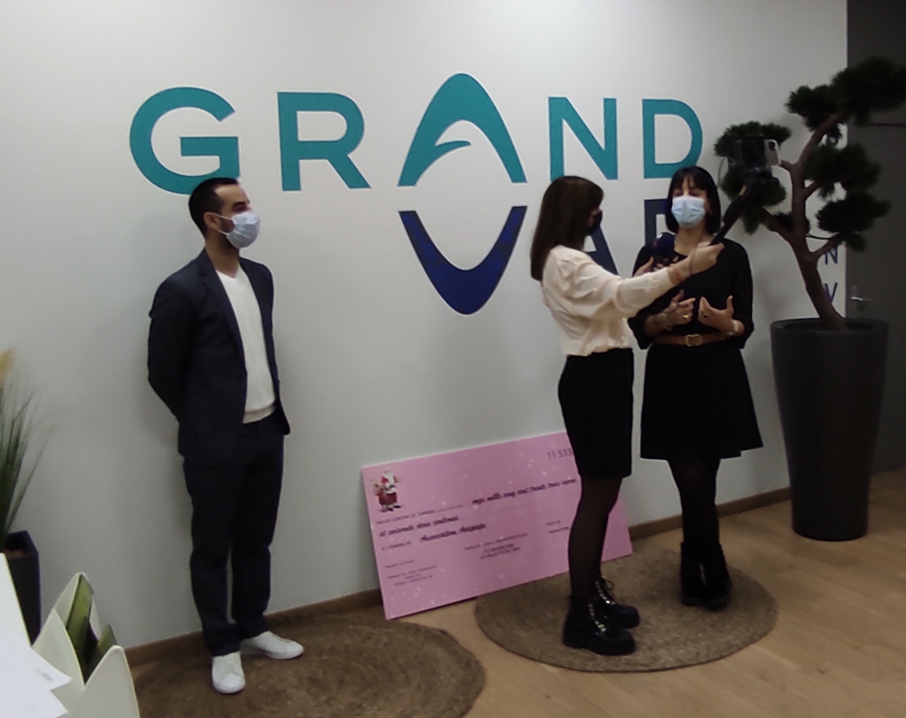 Entretien au micro devant le logo Grand Var, avec un grand chèque rose au sol.