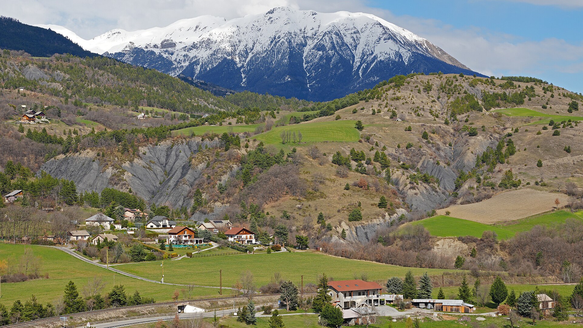 Vue de la vallée de la Durance dans les Hautes-Alpes, avec un sommet enneigé dominant des prairies et un hameau.