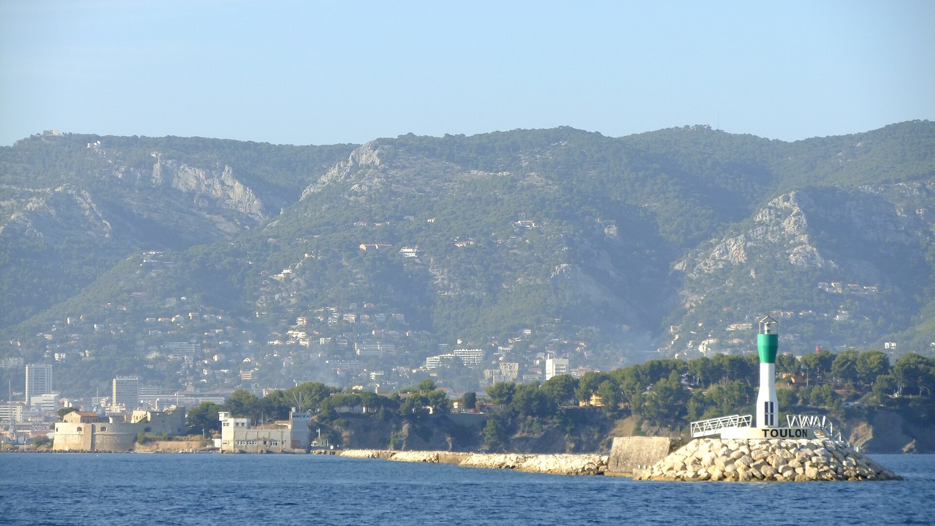 Le feu vert et blanc de la digue marquant l'entrée de la rade de Toulon, avec la ville et les collines du Var en arrière-plan.