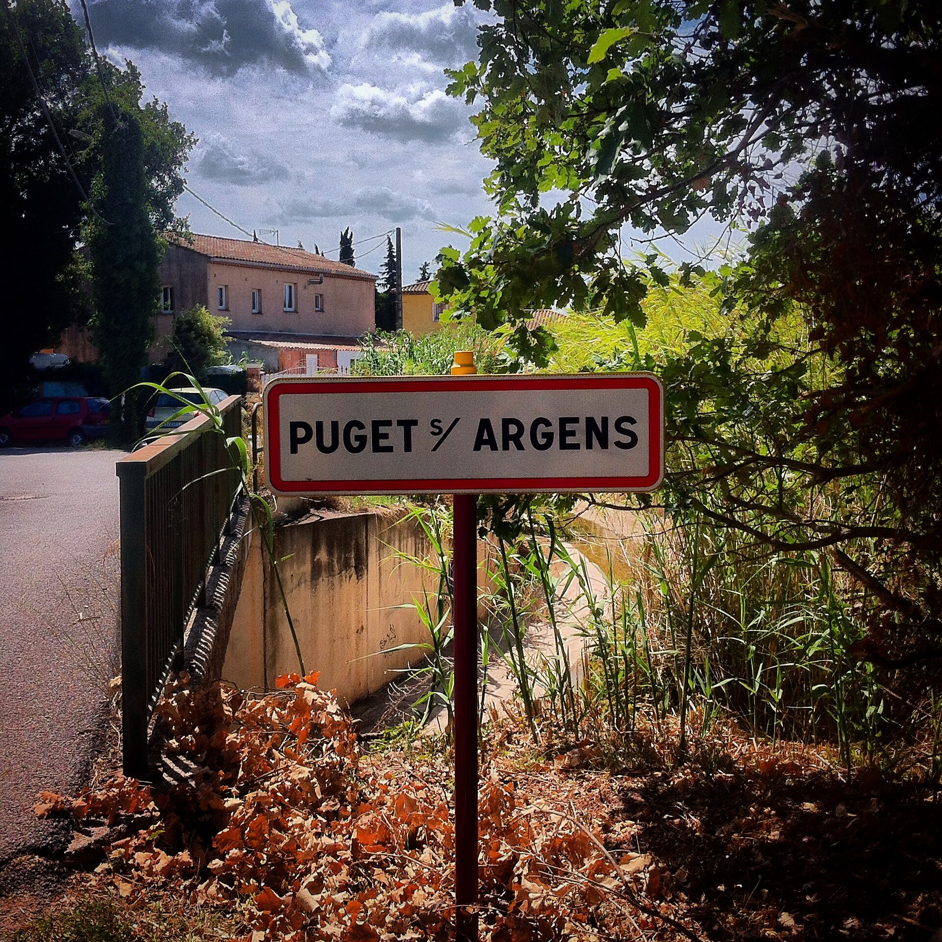 Panneau de Puget-sur-Argens au bord d’une route ombragée.